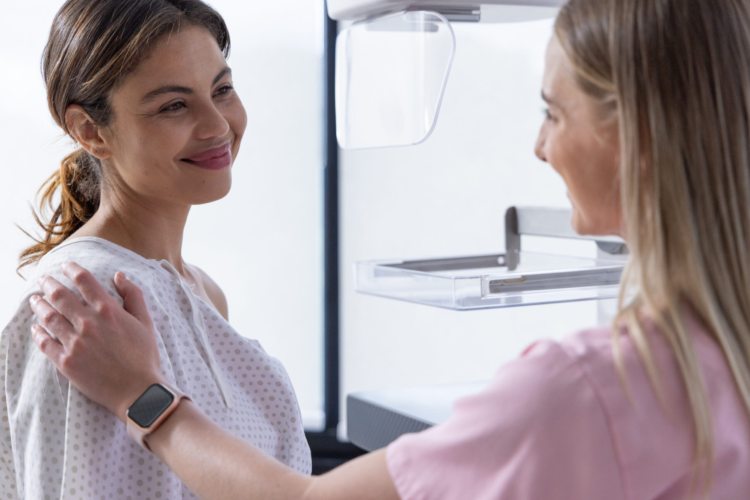 Female patient and tech in pink scrubs preparing mammogram in clinic, gown, watch, tray, copy space